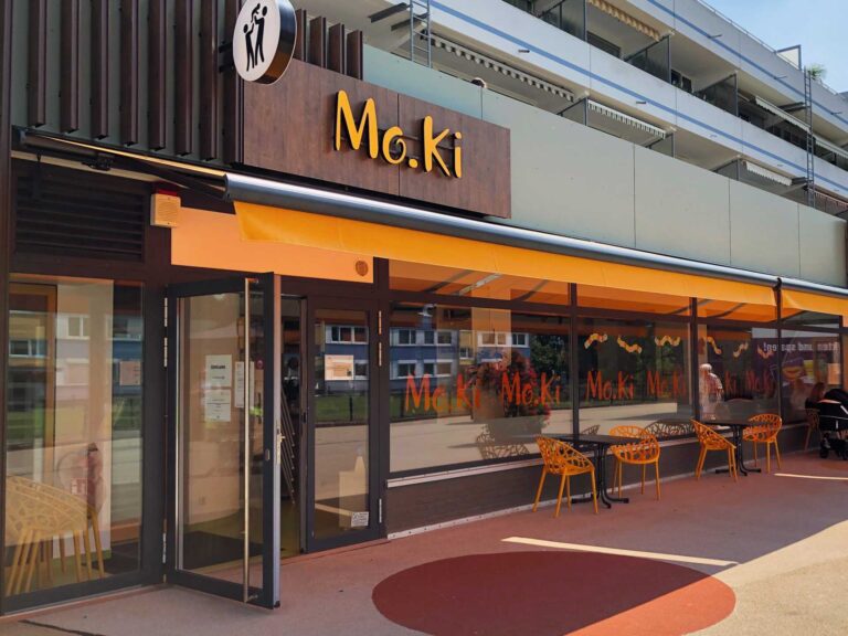 Außenansicht des Moki-Cafés in Baumberg mit Logo und Icon über dem Eingang und orangenen Stühlen vor dem Schaufenster