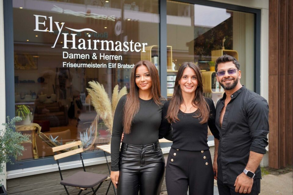 Die Friseurmeisterin und ihr Team (zwei weitere Personen) stehen gemeinsam vor dem Friseurladen. Dahinter sieht man das Schaufenster mit Schriftzug und einem kleinen Gartentisch davor mit Holzstühlen.